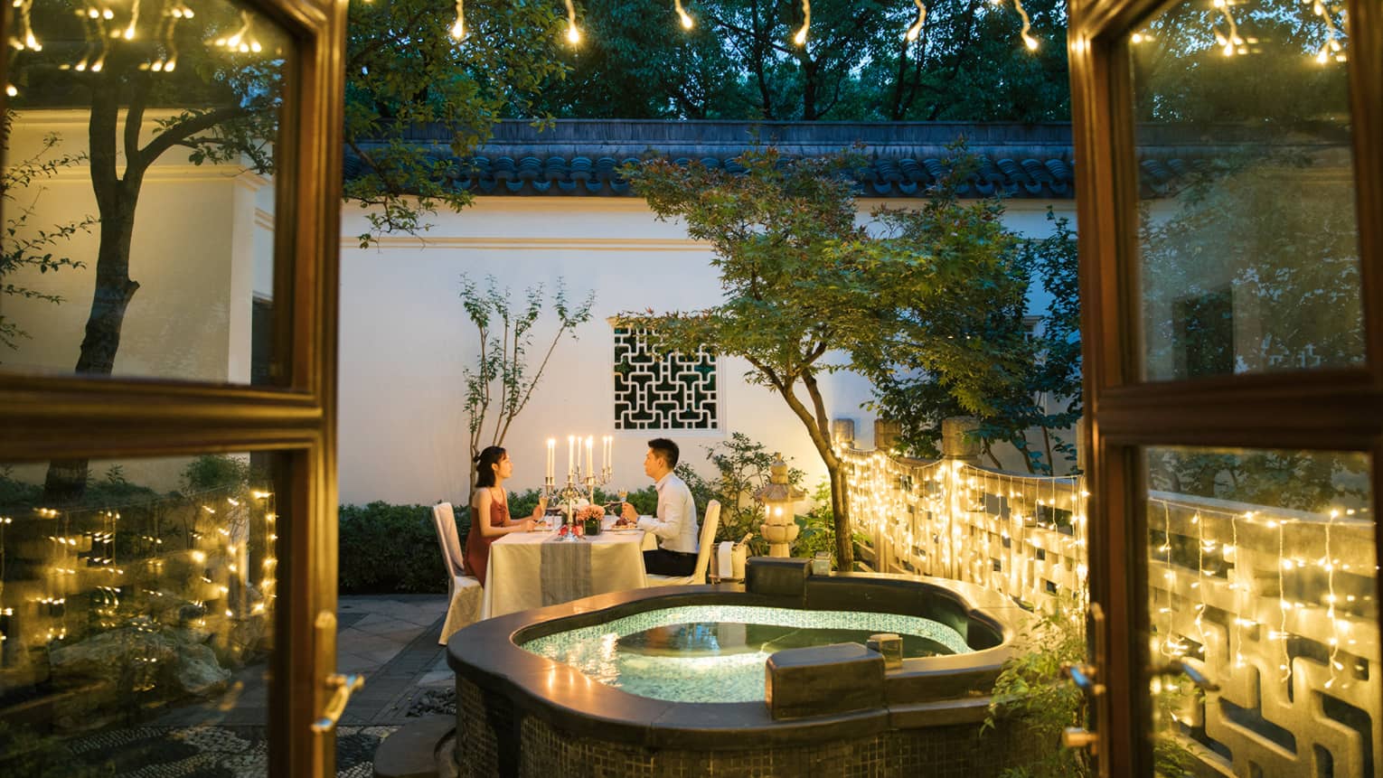 View past hot tub to couple dining at candlelit table on patio with lights