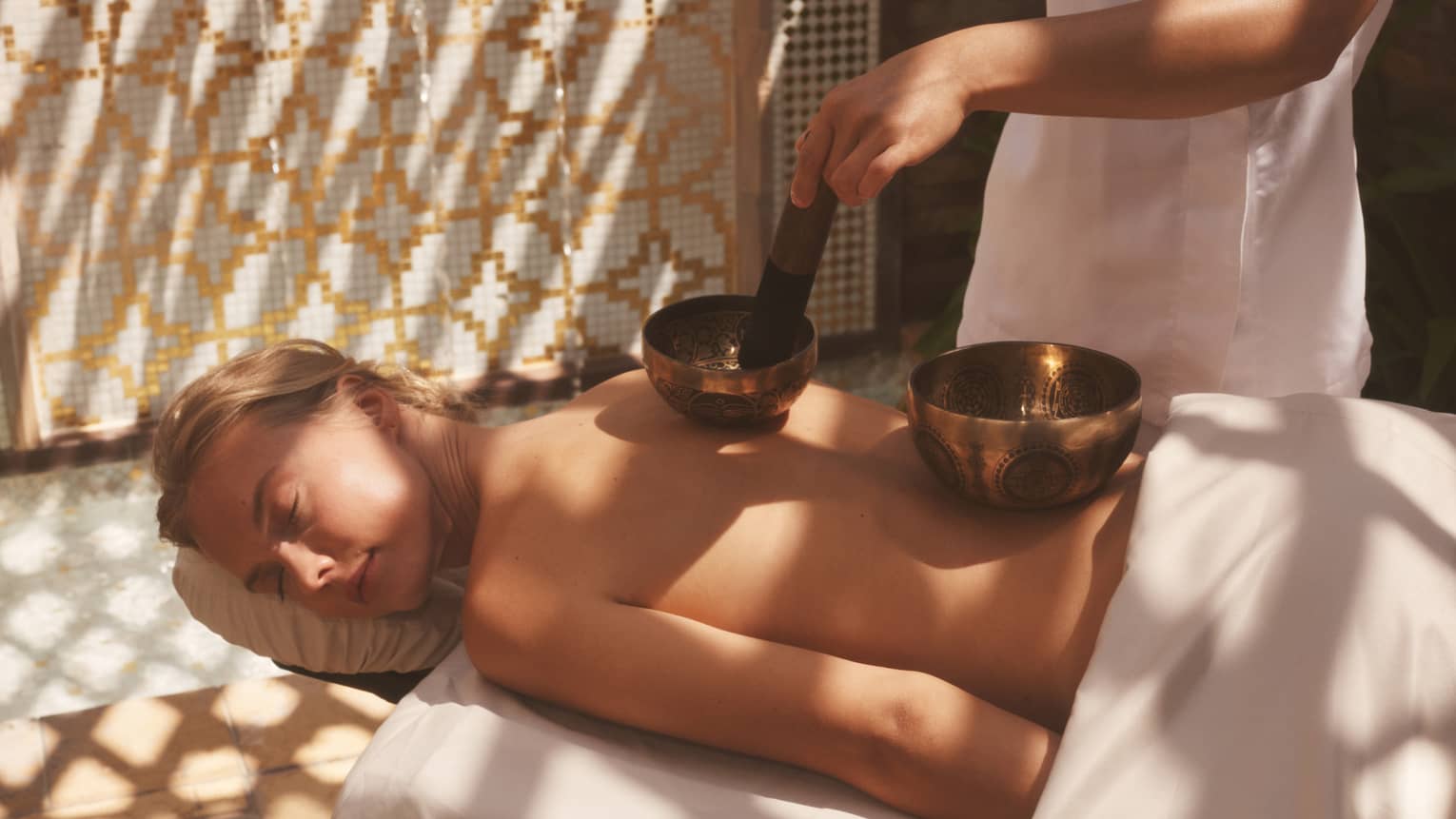 Unclothed person lying prone on a spa table with two sound healing bowls on back and spa therapist using them