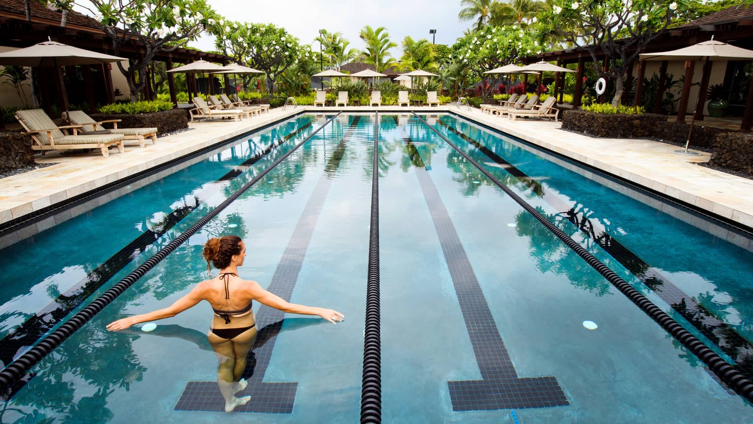 Back of woman in swimsuit wading in long outdoor swimming pool, arms outstretched