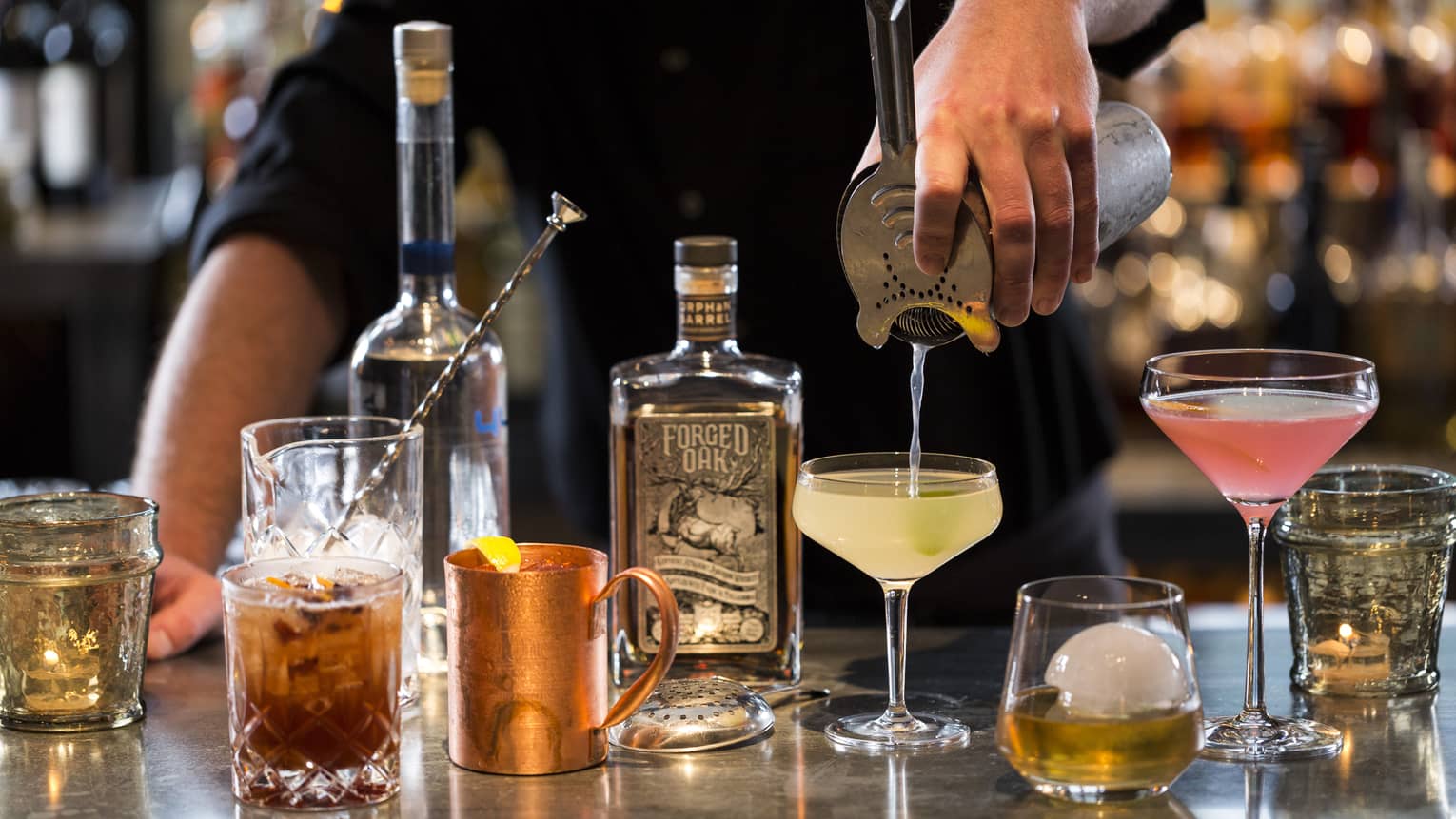 Various cocktail glasses and liquor bottles on bar as mixologist pours martini out of shaker