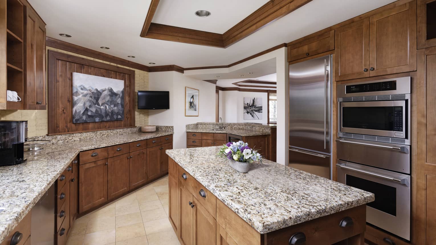 Luxury resort kitchen with dark wood cabinets, island, granite countertops, stainless steel appliances, TV and decorative artwork