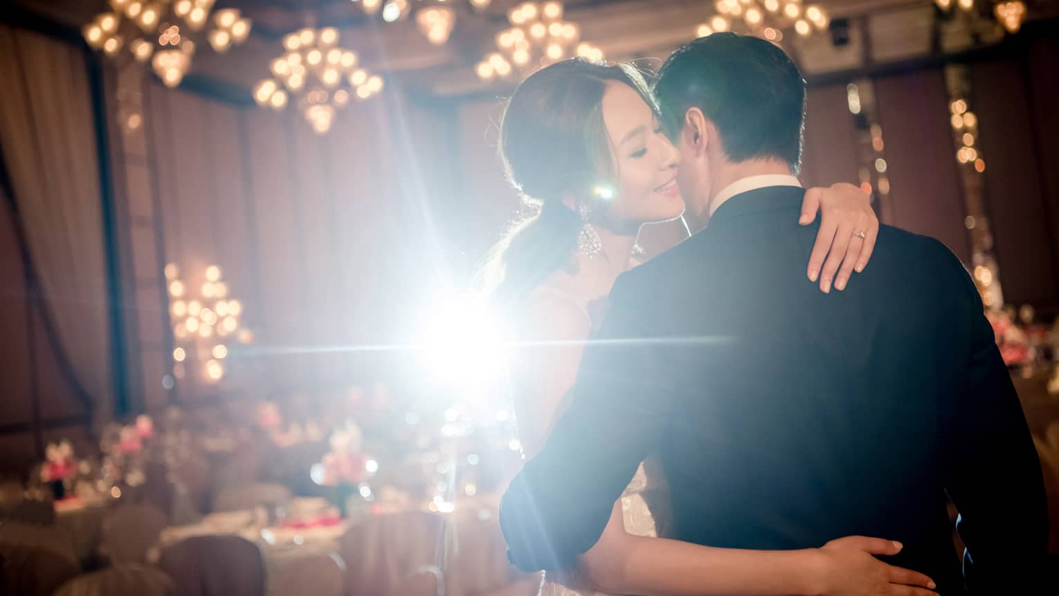 A bride and groom embrace each other in an indoor reception hall decorated with chandeliers and banquet tables