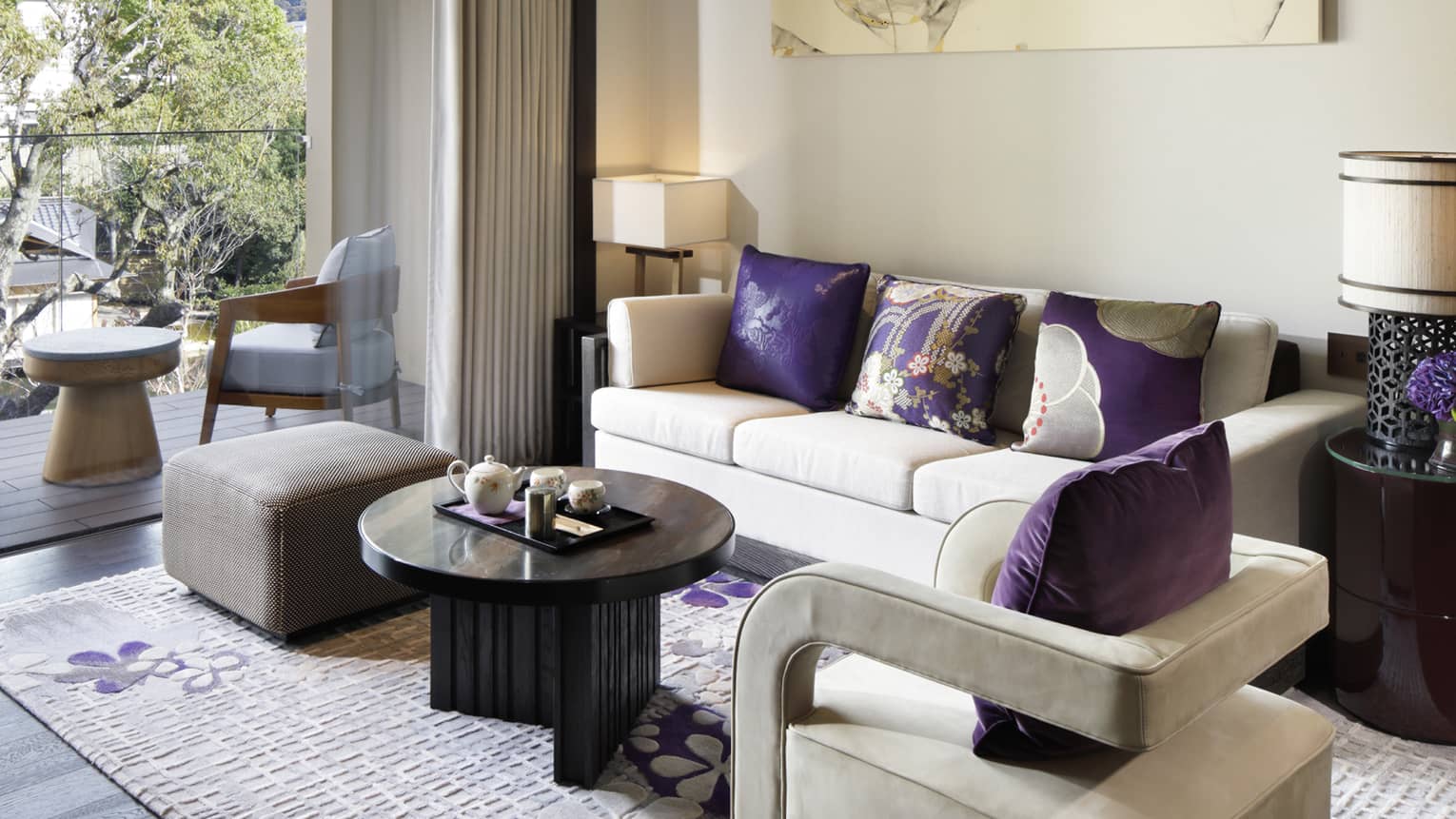 Living area with beige sofa, purple cushions, armchair, coffee table with tea set and balcony with outdoor seating and garden pond view