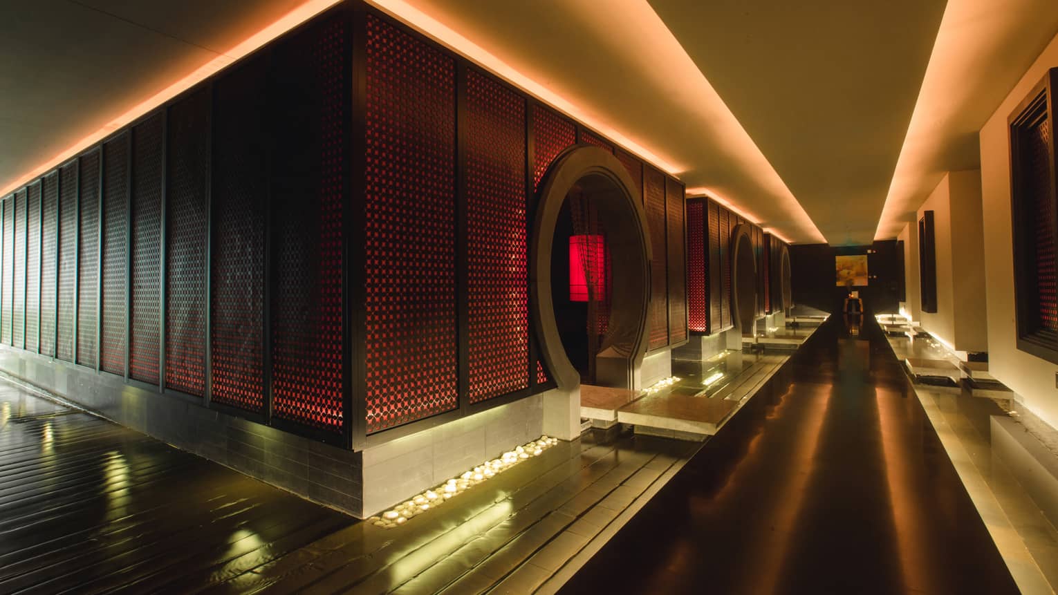 Dark floating walkway down dramatic spa hall past wall with red lights, circular doorways