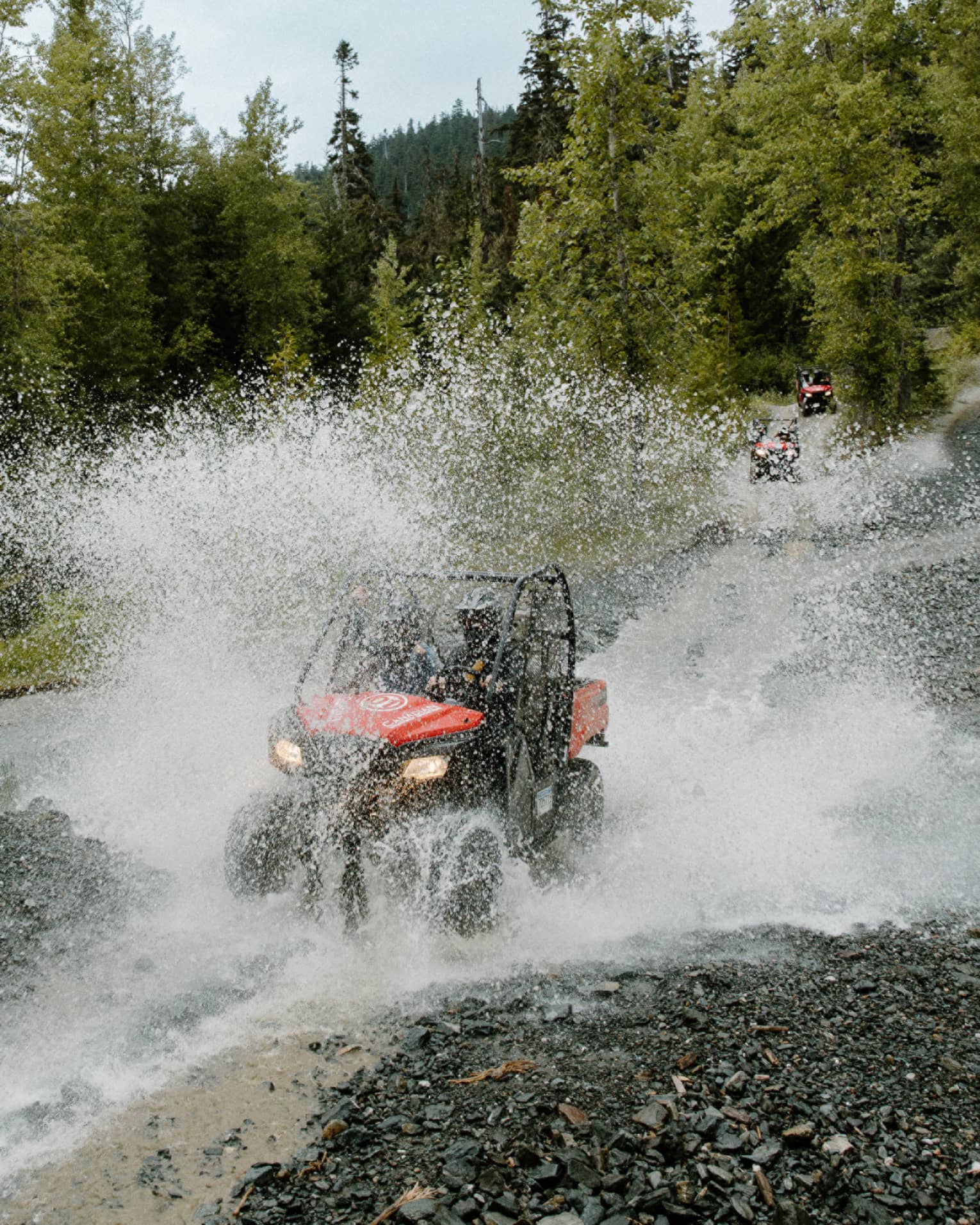 A red-and-black ATV driving through water, creating a thick veil of water spray against a background of pine forest.