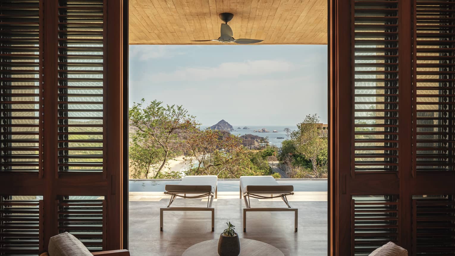 Open living area with wooden shutters framing a view of two lounge chairs on a terrace, overlooking an infinity pool, lush greenery and a distant ocean with rocky islands