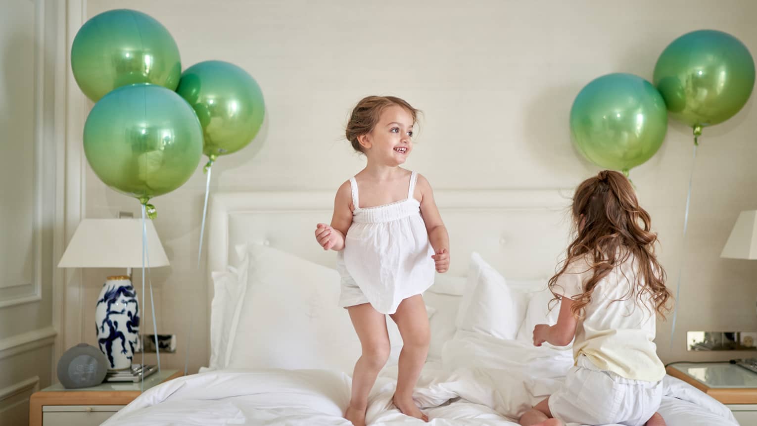 Two children jumping on white bed flanked with green balloons