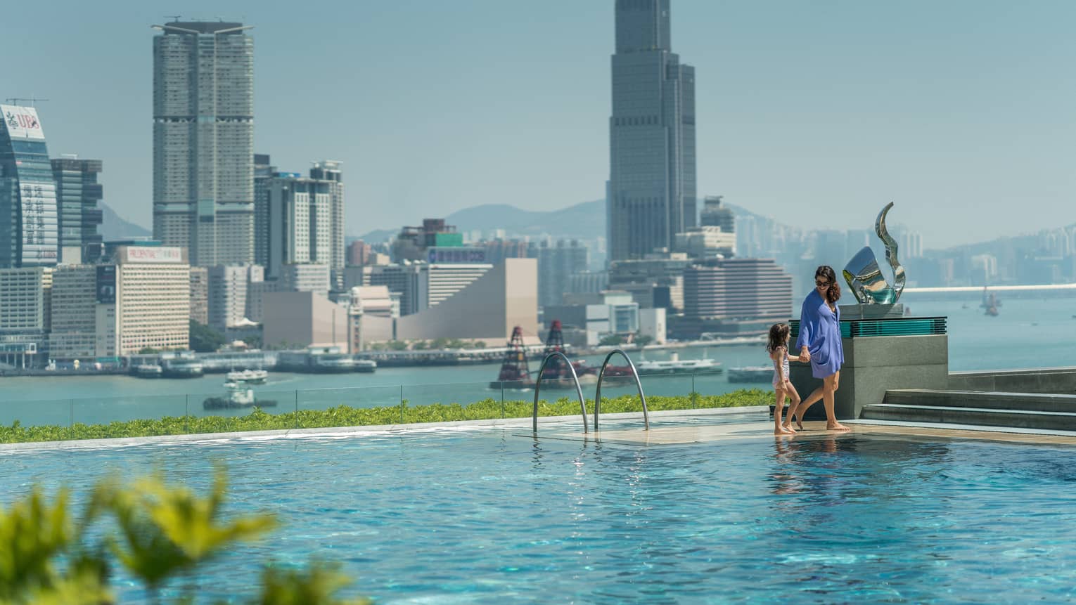 A parent and child walking along the edge of an infinity pool overlooking a skyline