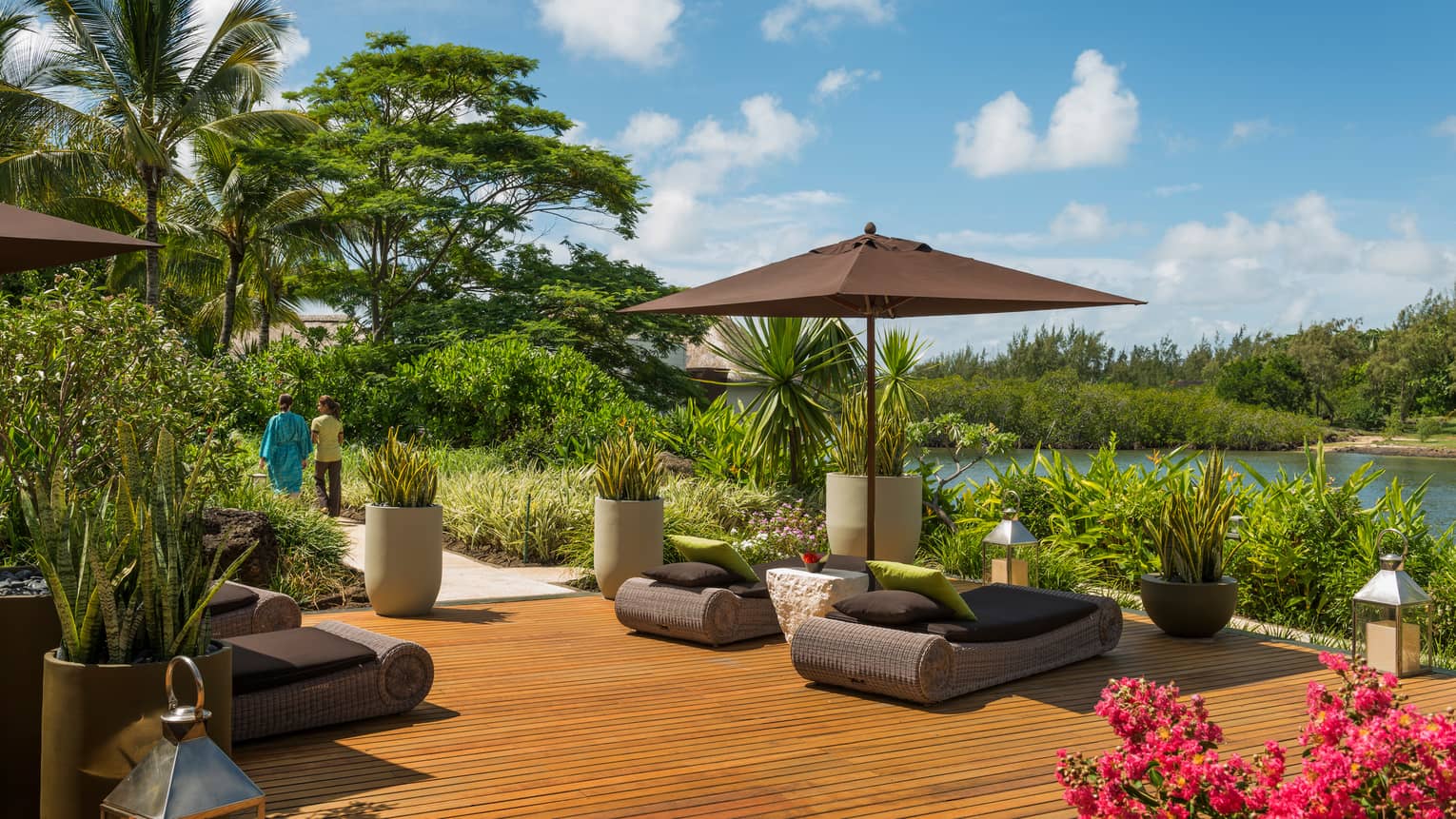 Woman in silk robe and hotel staff walk on path from wicker lounge beds on relaxation patio