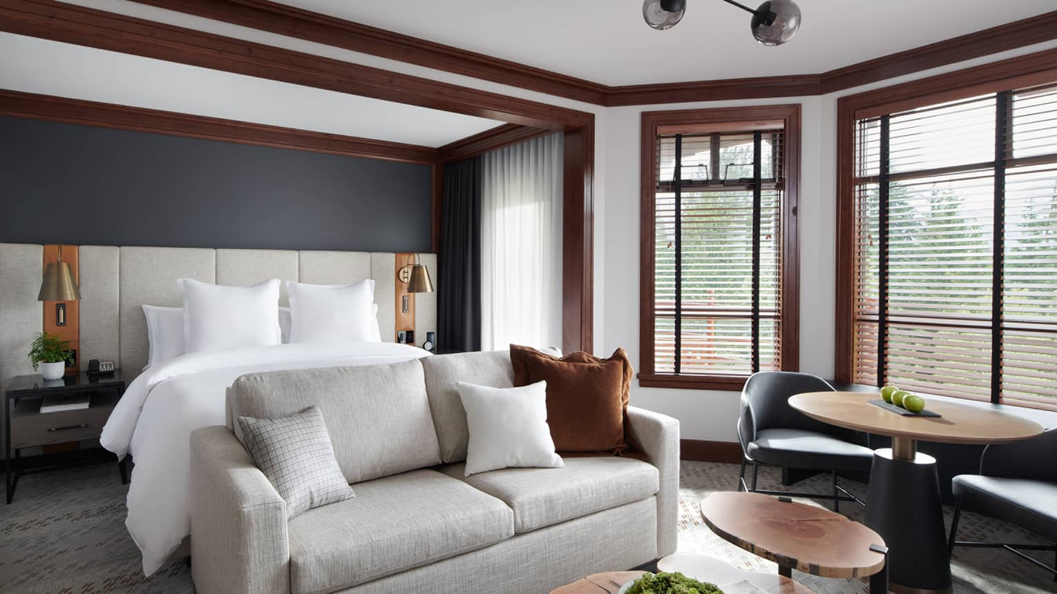 Elegant room at Four Seasons Whistler, showcasing a seamless blend of comfort and style. The room features a large, plush bed with crisp white bedding in the background, flanked by stylish bedside lamps. In the foreground, a cozy seating area includes a soft gray sofa with brown cushions, a circular table with chairs, and wooden coffee tables. Large windows with wooden blinds allow for natural light, enhancing the warm and welcoming atmosphere.,Elegant room at Four Seasons Whistler, showcasing a seamless blend of comfort and style. The room features a large, plush bed with crisp white bedding in the background, flanked by stylish bedside lamps. In the foreground, a cozy seating area includes a soft gray sofa with brown cushions, a circular table with chairs, and wooden coffee tables. Large windows with wooden blinds allow for natural light, enhancing the warm and welcoming atmosphere.