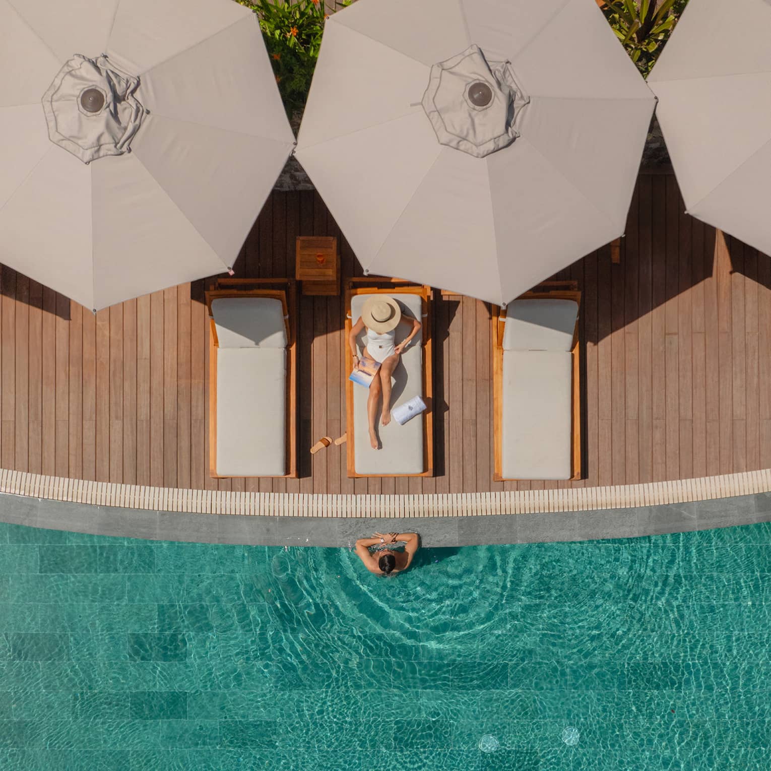 Aerial view of woman wearing white bathing suit relaxing on a lounge chair next to a pool with a man standing in the water with his arms on the ledge