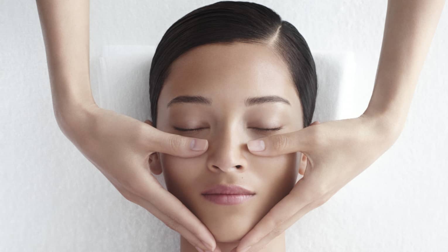Woman in white bathrobe lies on massage table with eyes closed, masseuse rests hands on her cheeks