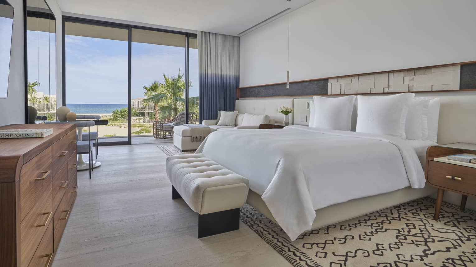 White bed with upholstered foot bench, side tables, dresser, lounge chair, tall glass doors and water views