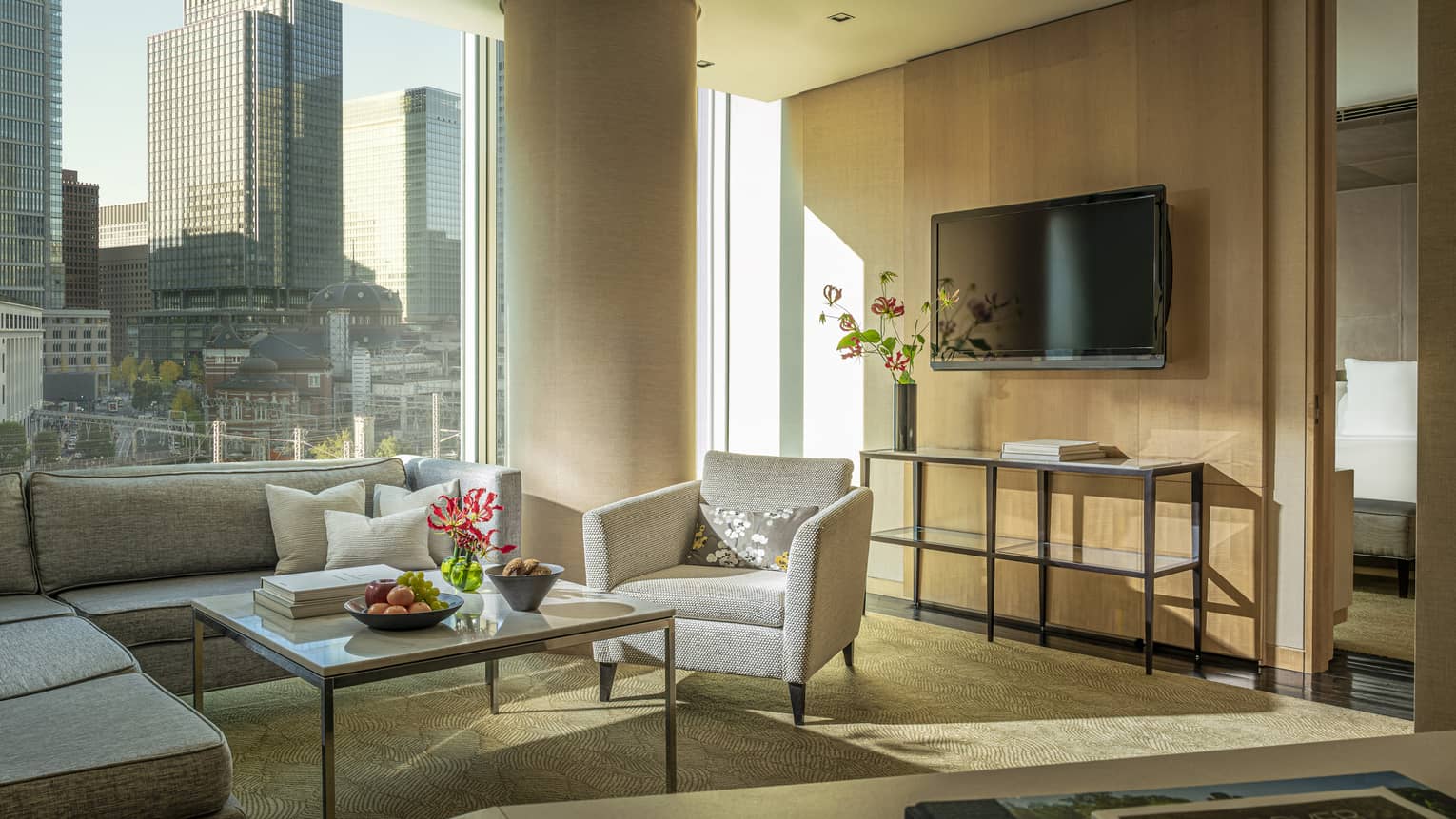 Four Seasons executive suite with gray sectional sofa, armchair, coffee table with fruit and flowers, large window with city view, TV and console table