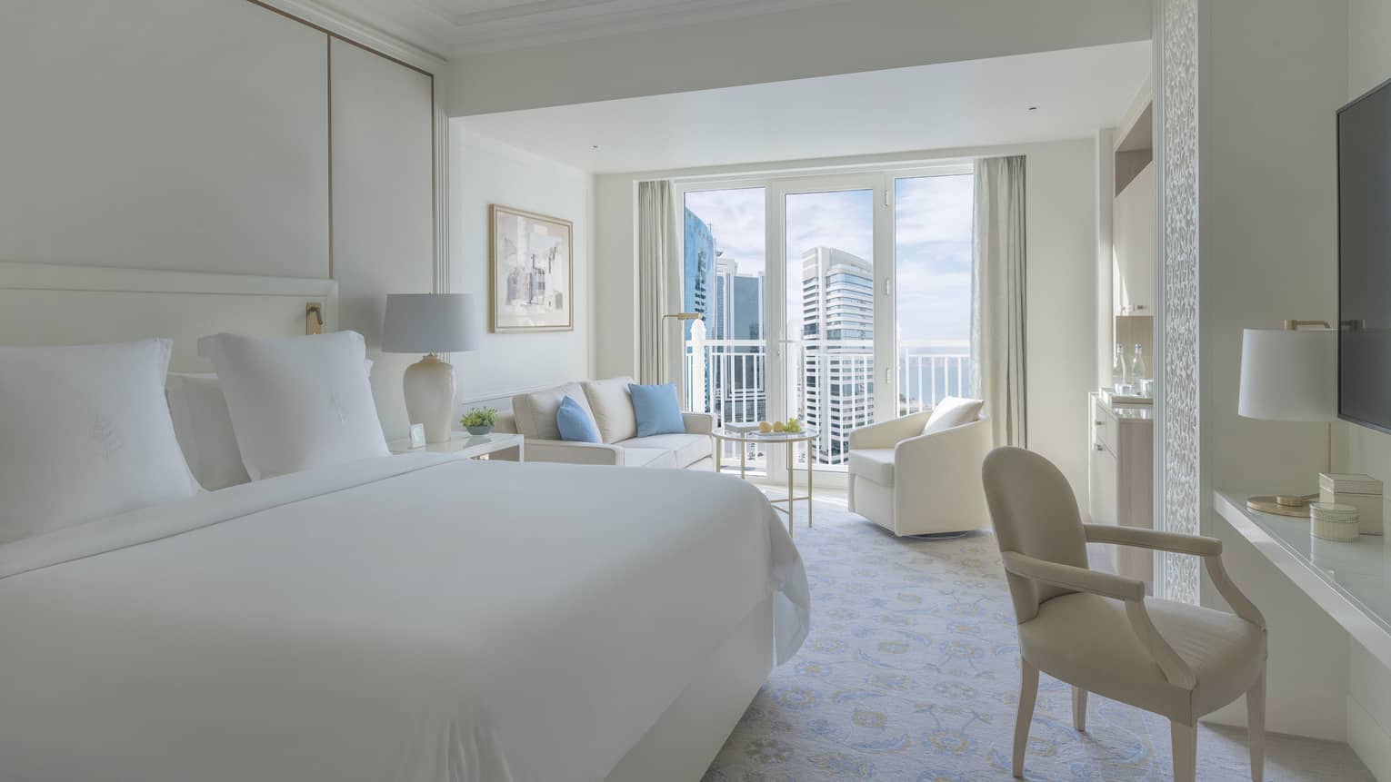Bedroom with white bed, white walls and a white sofa and arm chair, with balcony offering city view
