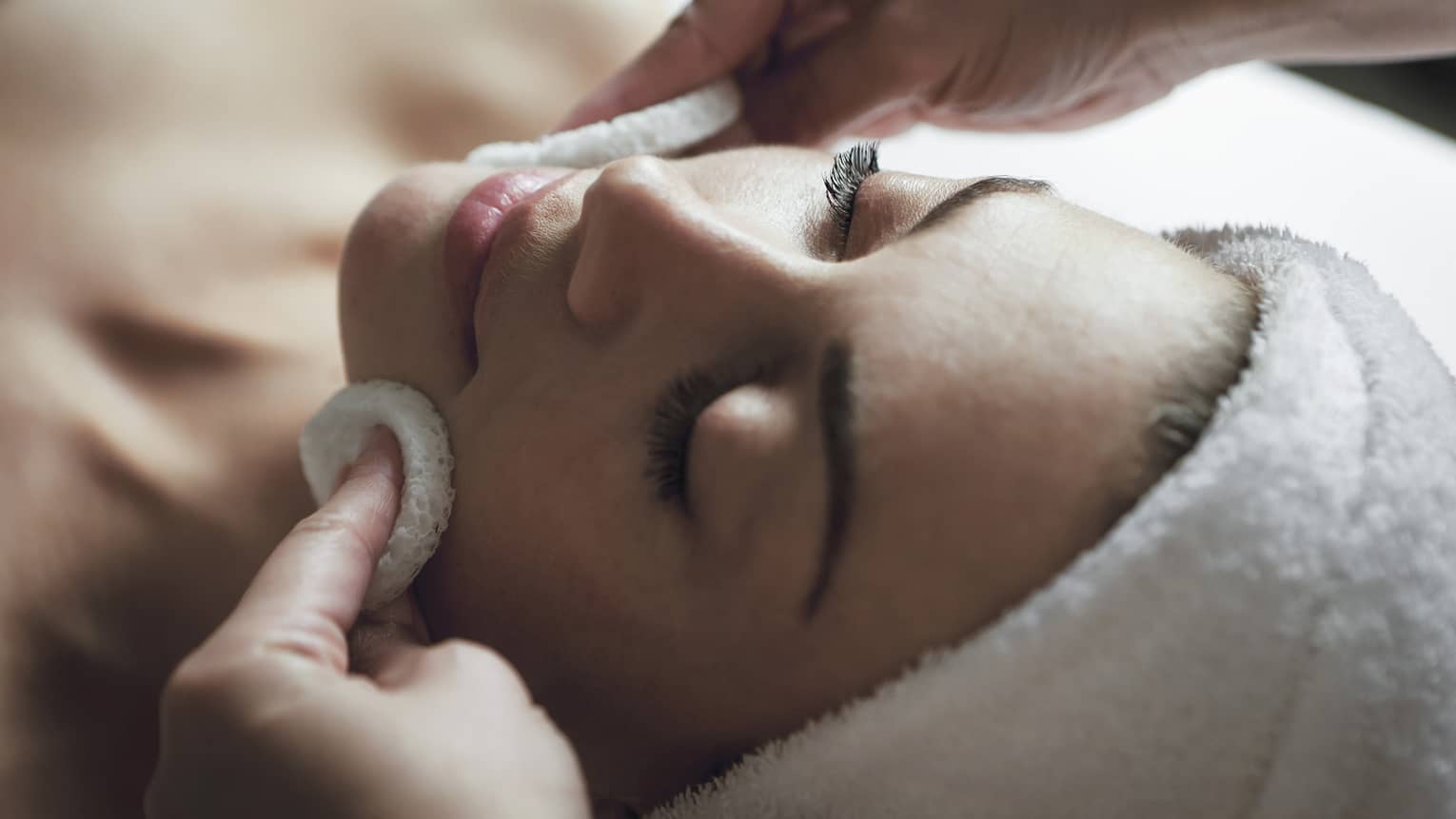 A detail of hands rubbing a sponge on a woman's face as she lies on a massage table with her eyes closed and hair wrapped up in a white towel