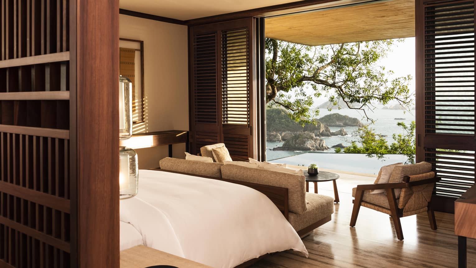 A serene hotel bedroom with wooden accents and a cozy seating area, opening to a terrace with breathtaking views of the ocean, rocky islands and lush greenery