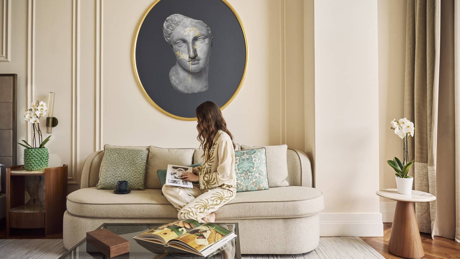 Guest with long dark hair and silk pajamas sits on a cream loveseat reading a magazine with a large round painting of an ancient Roman bust hanging on the wall behind her