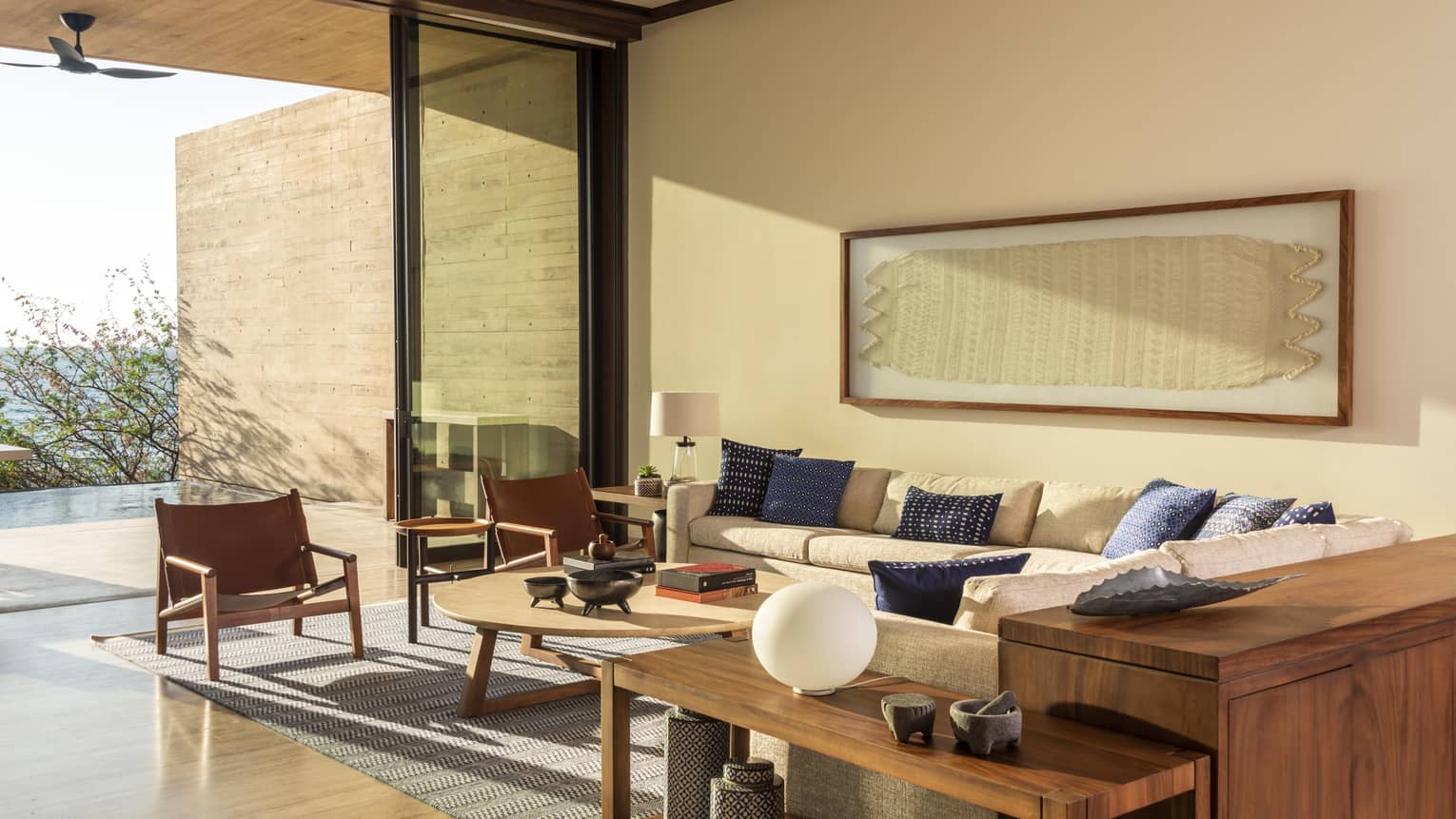 Modern living room with a beige sectional sofa, blue accent pillows and wooden furniture. Large glass doors open to a patio, allowing natural light to fill the space.