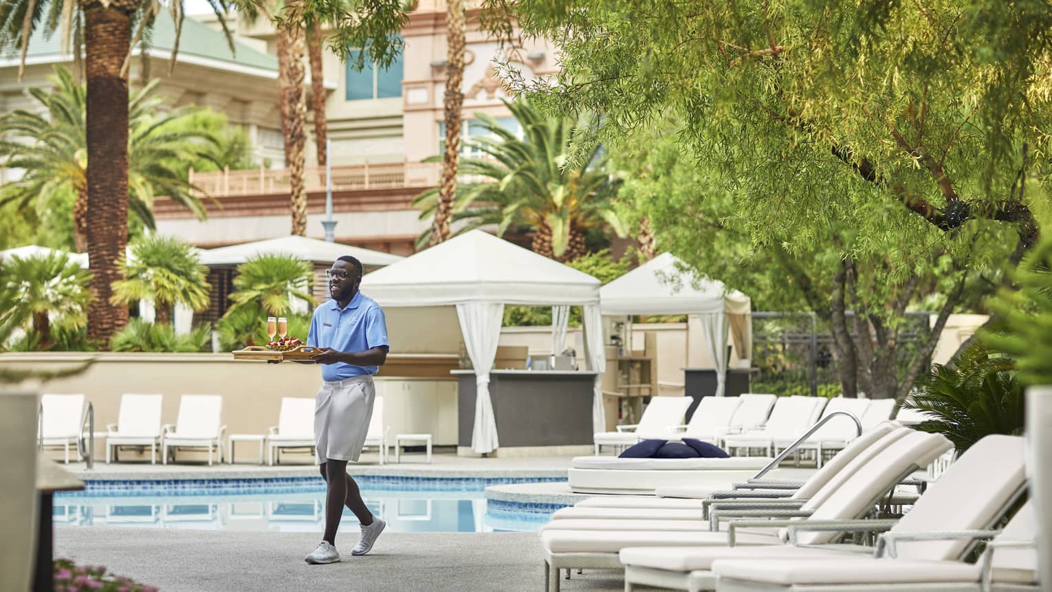 Man in blue shirt and kaki shorts walking with tray of drinks by outdoor pool
