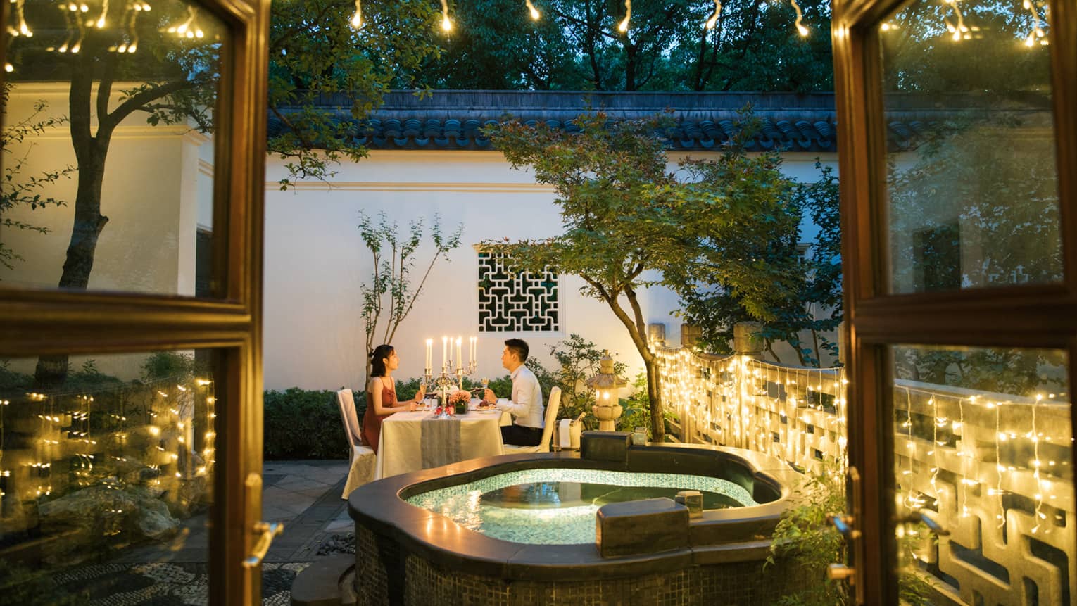 View past hot tub to couple dining at candlelit table on patio with lights