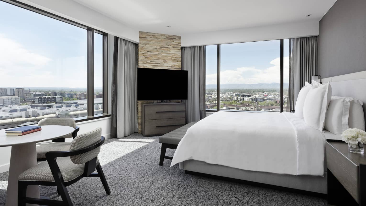 A hotel room with a white sheet covered bed, a TV, small white table with grey chair and large windows on two walls.