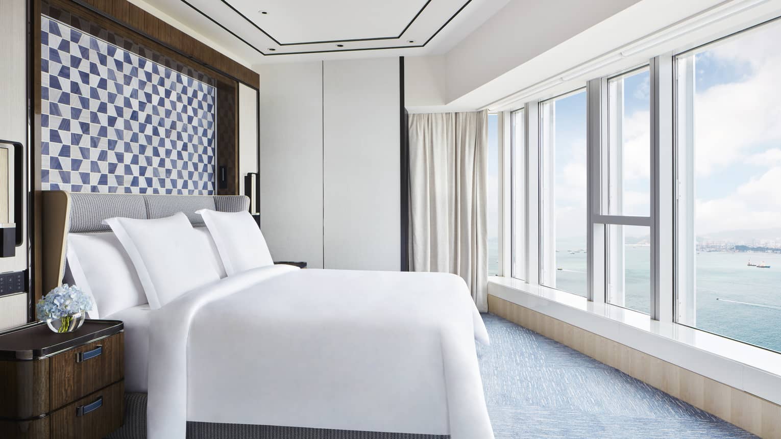Bright hotel bedroom with large bed, blue geometric headboard, floor-to-ceiling windows and ocean view.