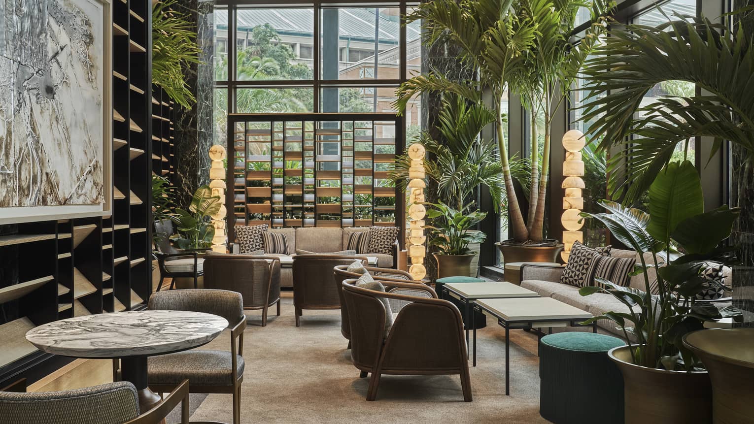Chandelier Bar seating area with brown accent chairs, marble-topped tables, floor-to-ceiling windows and potted palms