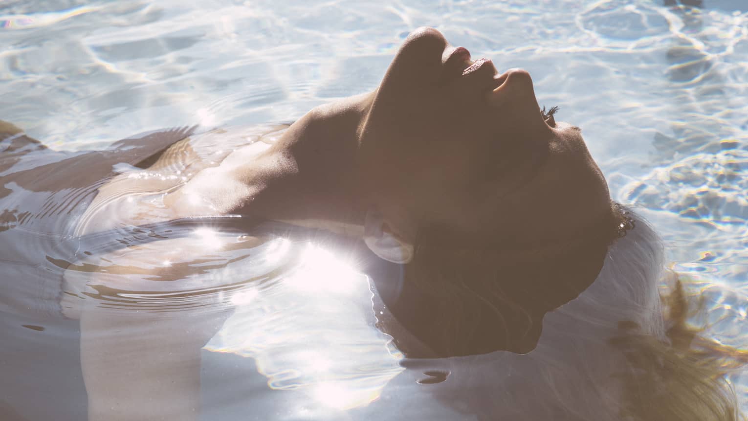 Close-up of woman's head as she floats on her back in pool, sunlight reflecting, her hair flowing in water