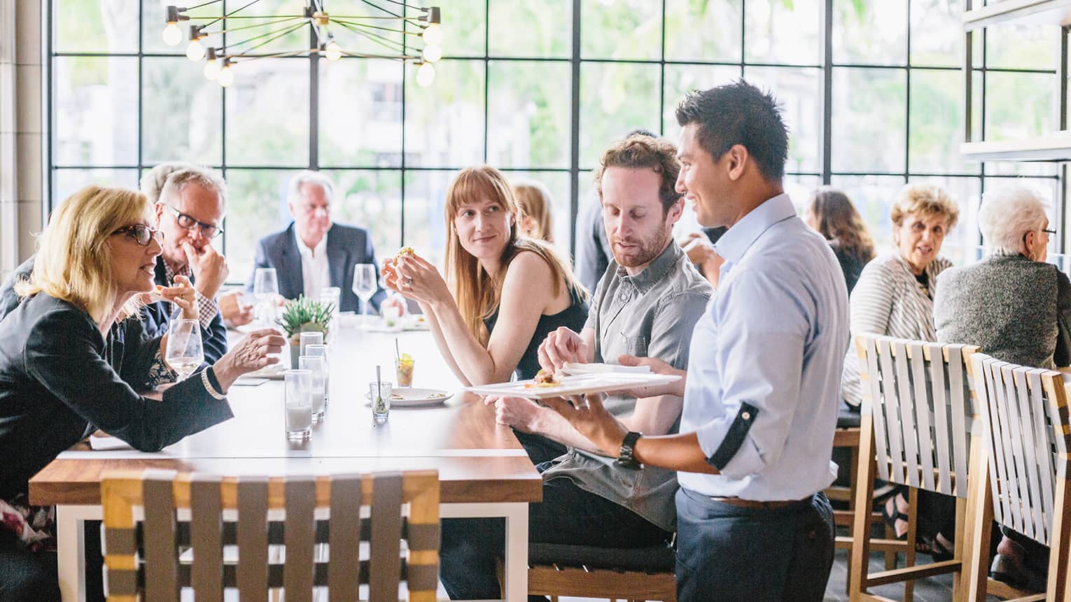 Crowds gathered around long dining table in sunny Seasons Restaurant as server stands with tray of appetizers