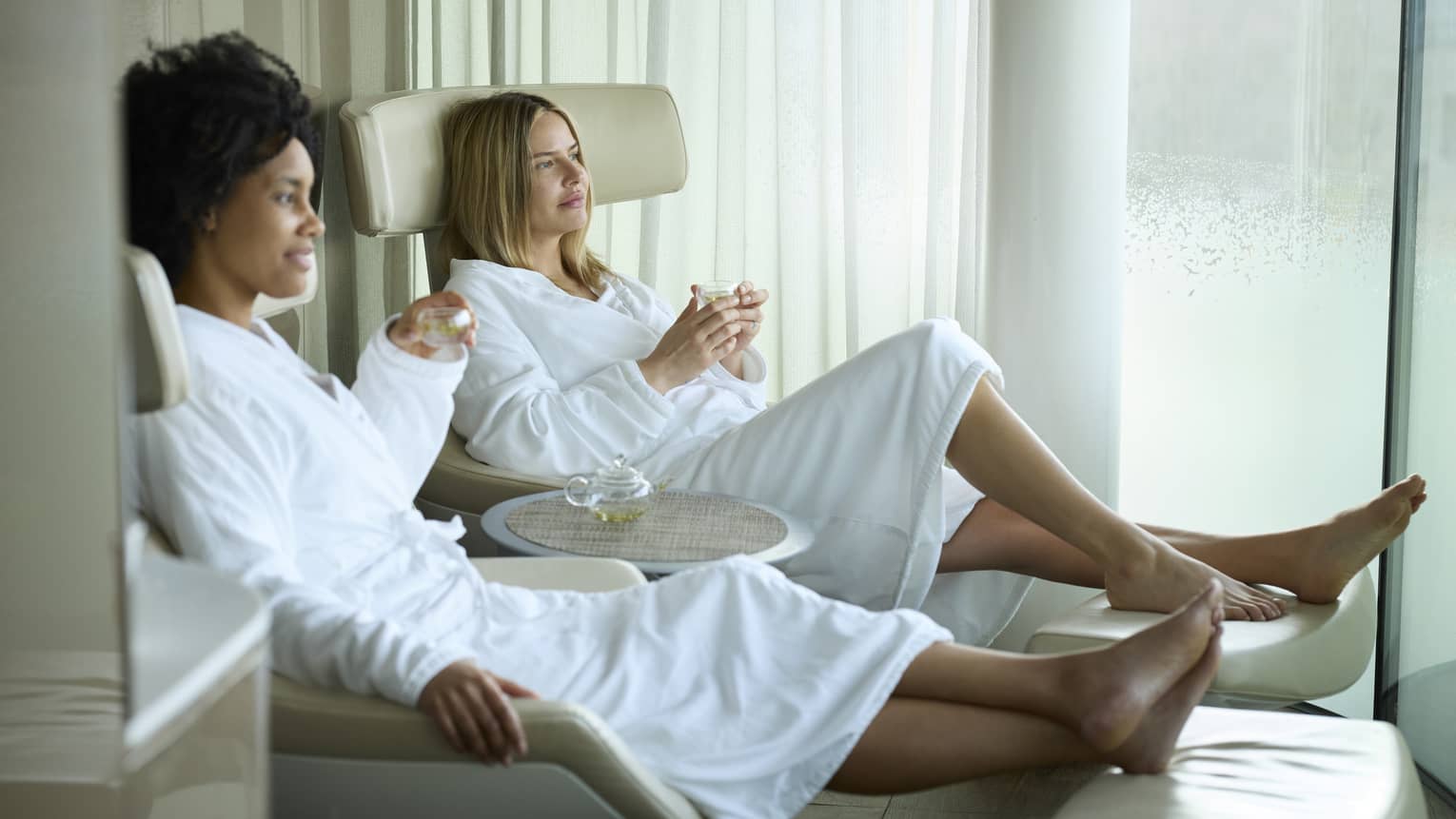 Two spa guests in white robes sitting on lounge chairs drinking champagne