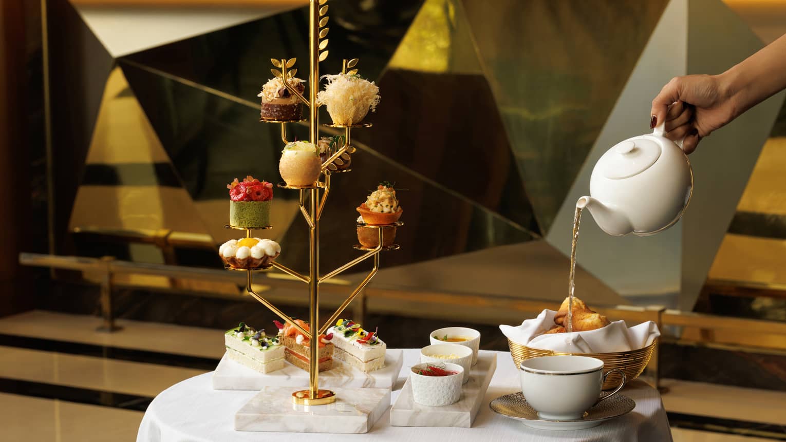Table set for afternoon tea with a gold tree-shaped pastry stand, white marble platters with finger sandwiches and small bowls, a gold break basket and a hand reaching in pouring tea from a white teapot into a white mug