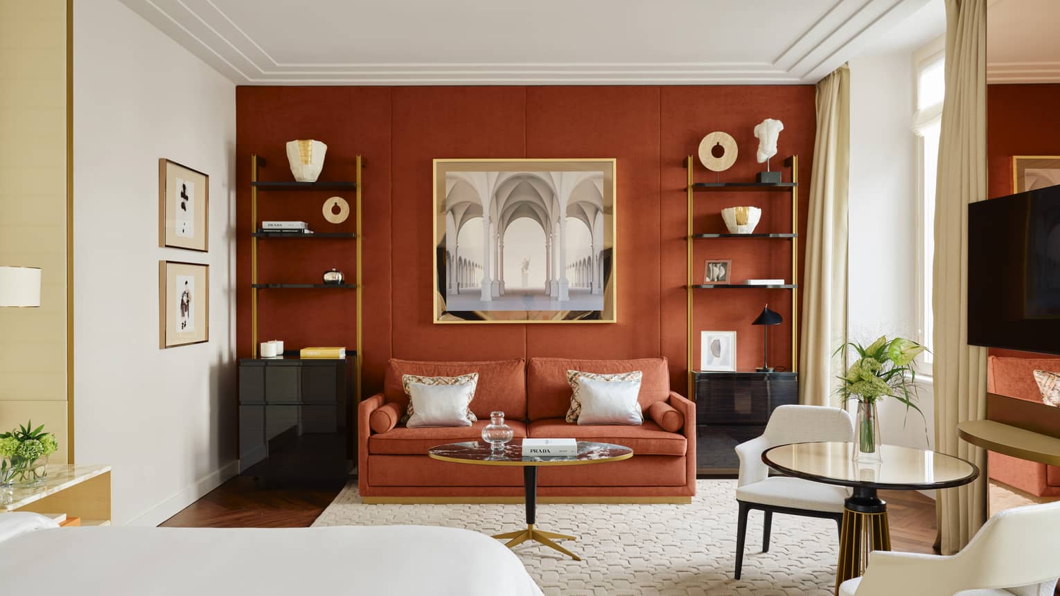 Contemporary hotel room with a dark burnt-orange accent wall and matching coloured sofa, two shelves containing artwork and crown molding