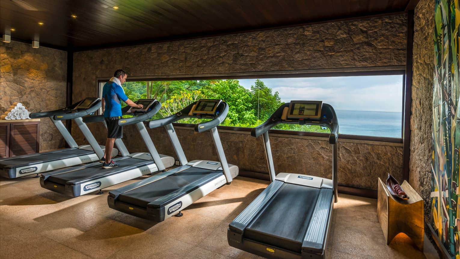 Man with white towel around shoulder walks on treadmill beside cardio machines in front of window
