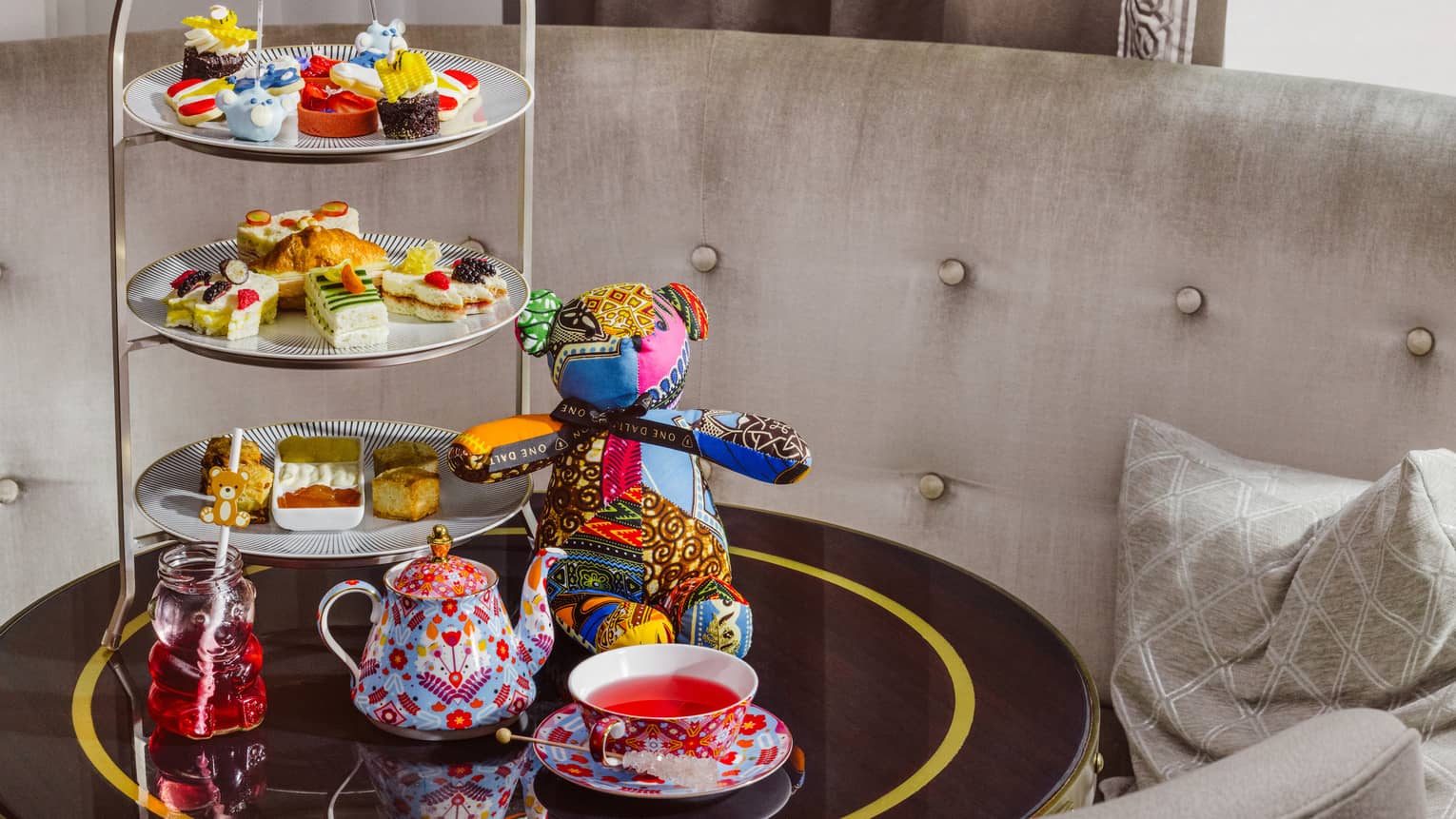 A small tabletop next to upholstered seating holds a decorative teapot and cup, a display of small bites, and a colourful stuffed teddy bear.