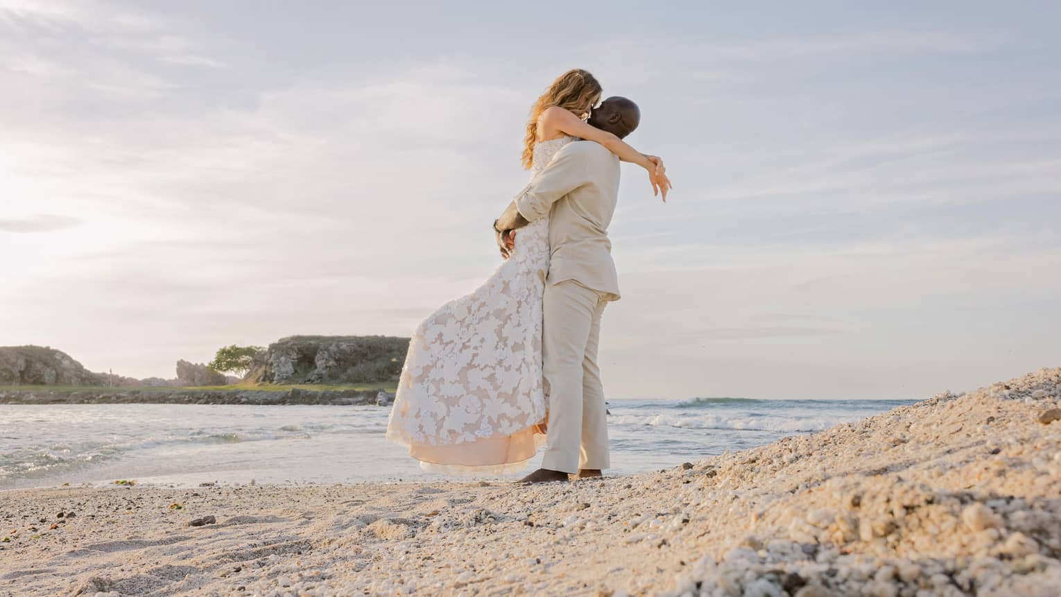 A bride and groom embrace on a beach at sunset