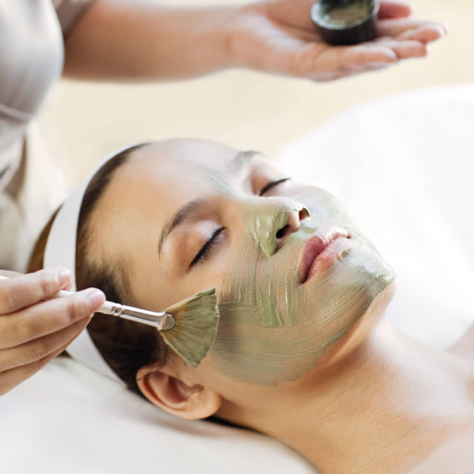 Spa staff holding brush paints green mask on woman's cheeks, chin, nose