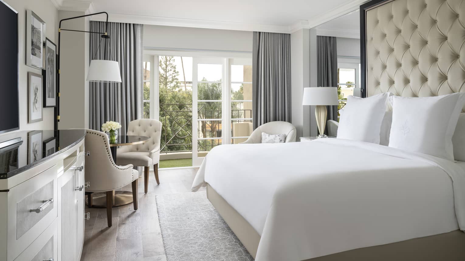 A hotel room with a large bed with white sheets, black counter on white dresser, three chairs, and a large window.
