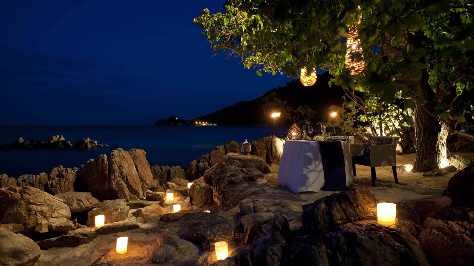 Candles glow on rocks near private dining table on beach at night