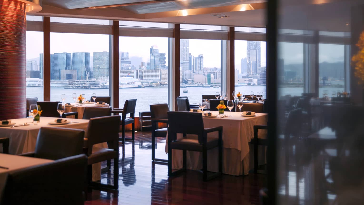 A sophisticated restaurant with neatly set tables and chairs. Large windows offer panoramic views of the city skyline and the waterway.