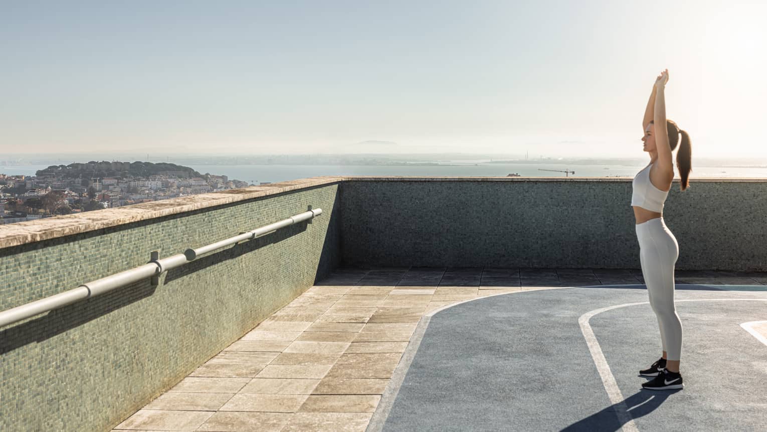 A person stretches with arms overhead while standing on a rooftop running track on a sunny day, with the horizon between water and sky visible in the background