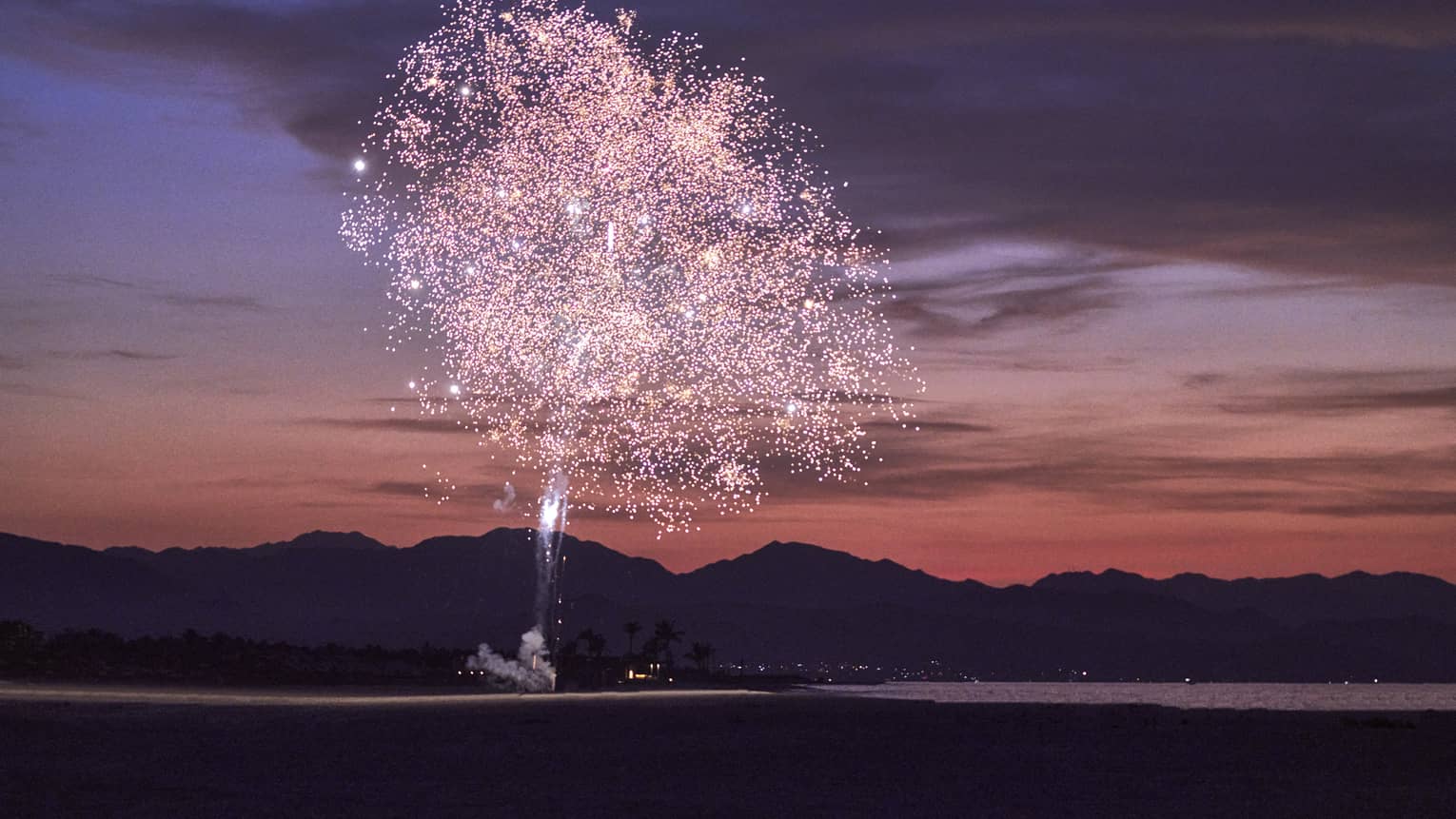 Pink fireworks exploding during sunset with a mountain behind them.