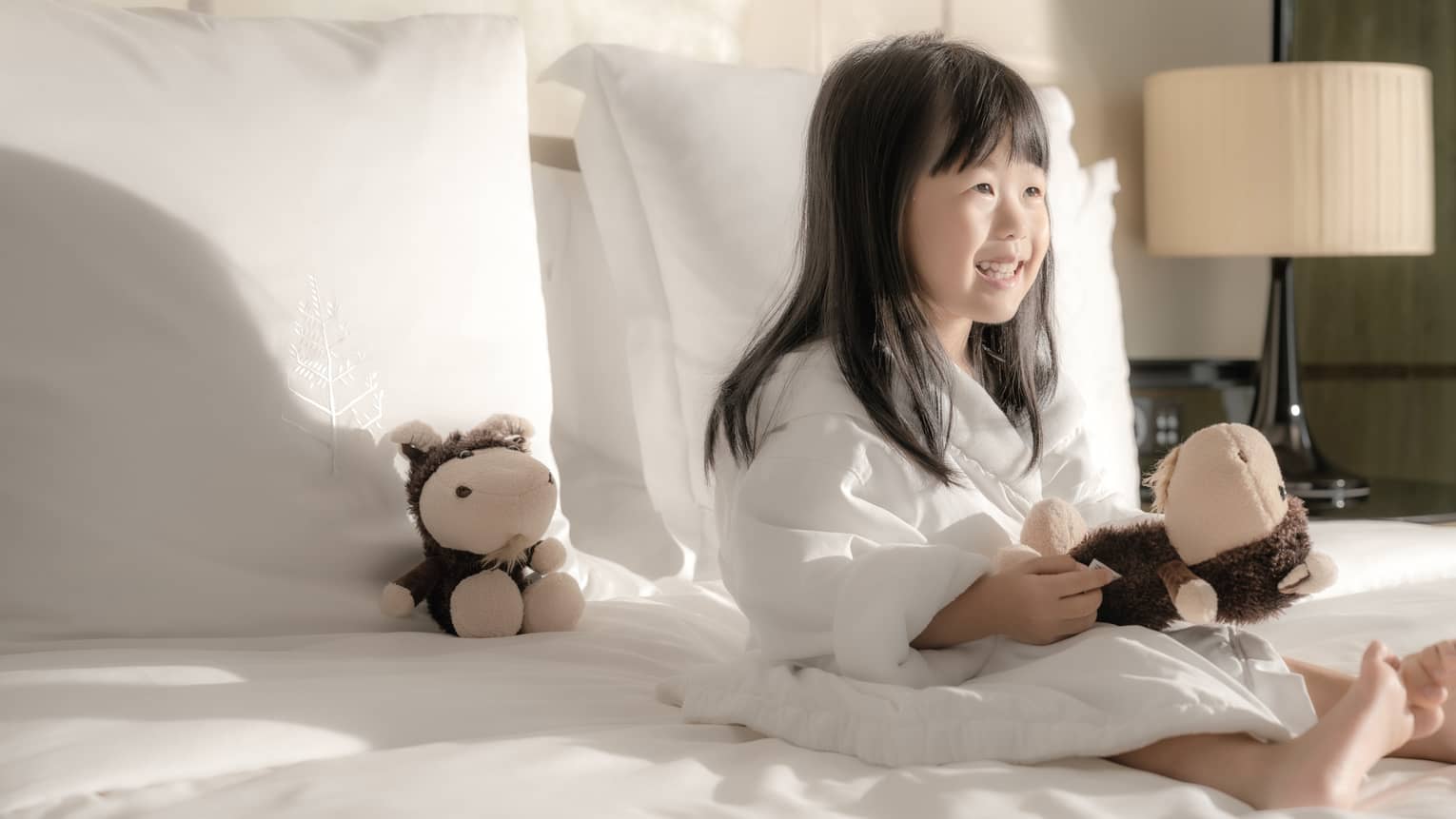 Smiling young girl in bathrobe holds stuffed animal on bed with white linens