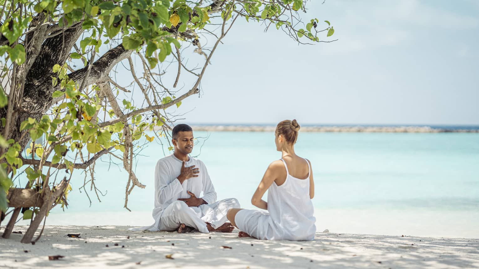 Dr. Arun K. Tomson practises breathing exercises with female guest on the beach under the shade of a tree