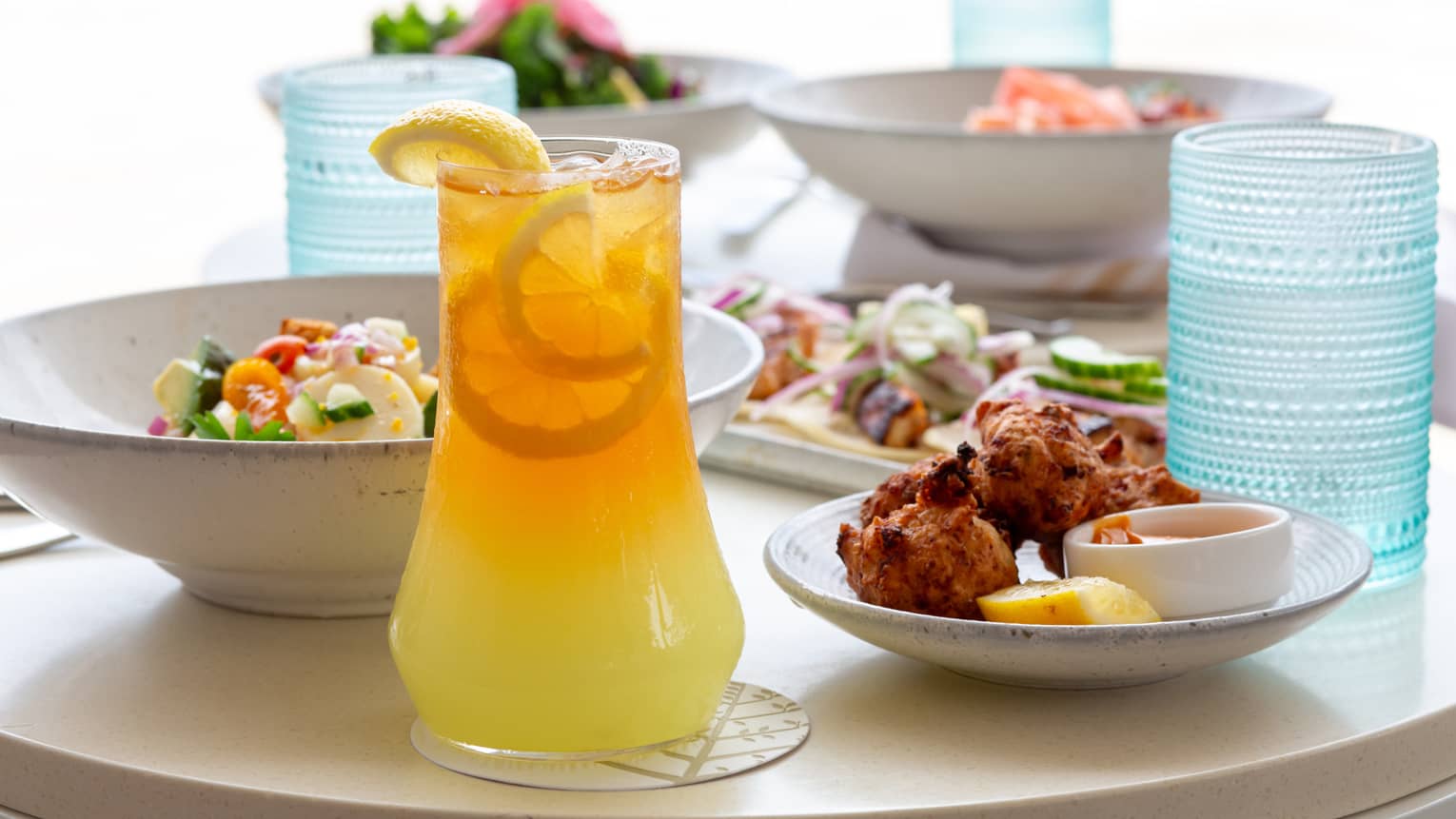 Large glass pitcher of orange juice, a small plate of chicken wings, a bowl of salad, a rectangular plate of tacos and two more bowls of food fill a white table
