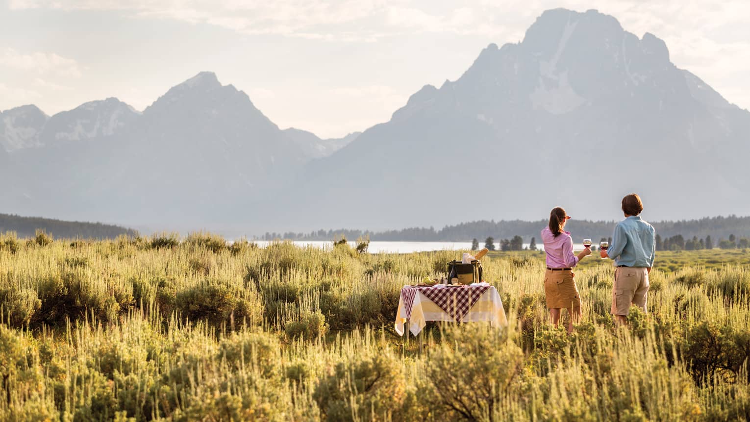 A man and woman sip wine in the grasslands while gazing out at the mountains at sunset