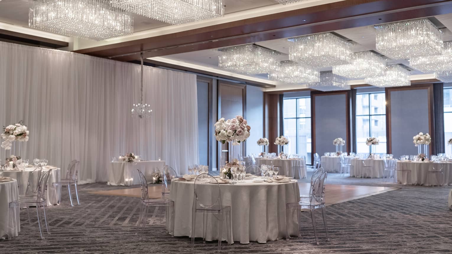 A ballroom with round tables with flower centerpieces surrounding a section of hardwood floor, many lights hang from the ceiling.