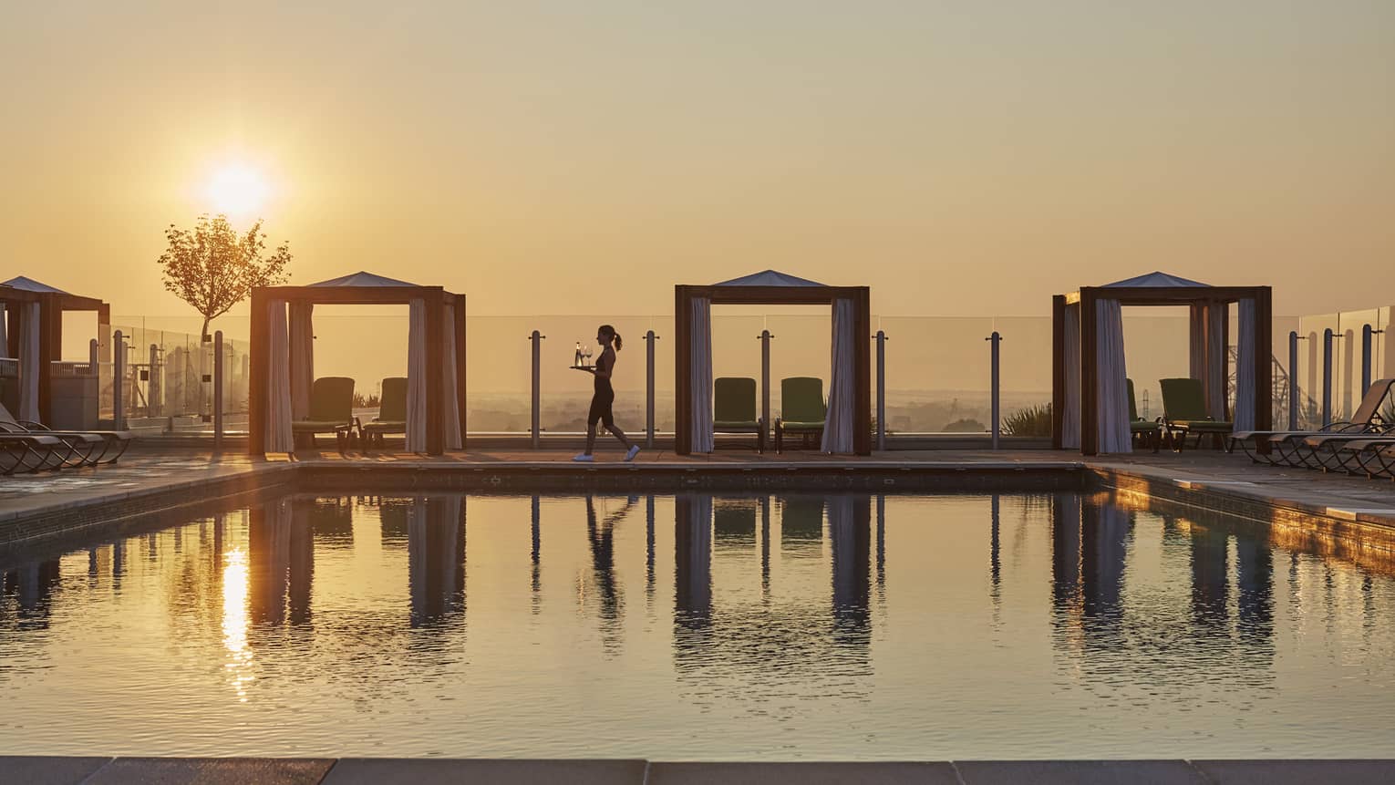 A server walking by a rooftop pool with cabanas at sunset and carrying drinks, with the golden sky reflecting on the water in a luxury hotel terrace setting