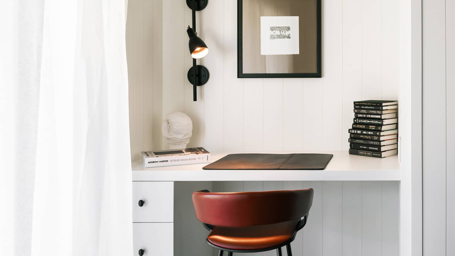 Close-up of a luxury resort room desk with black wall lamp, stacked books, art print, red leather chair and decorative bust on a white workspace