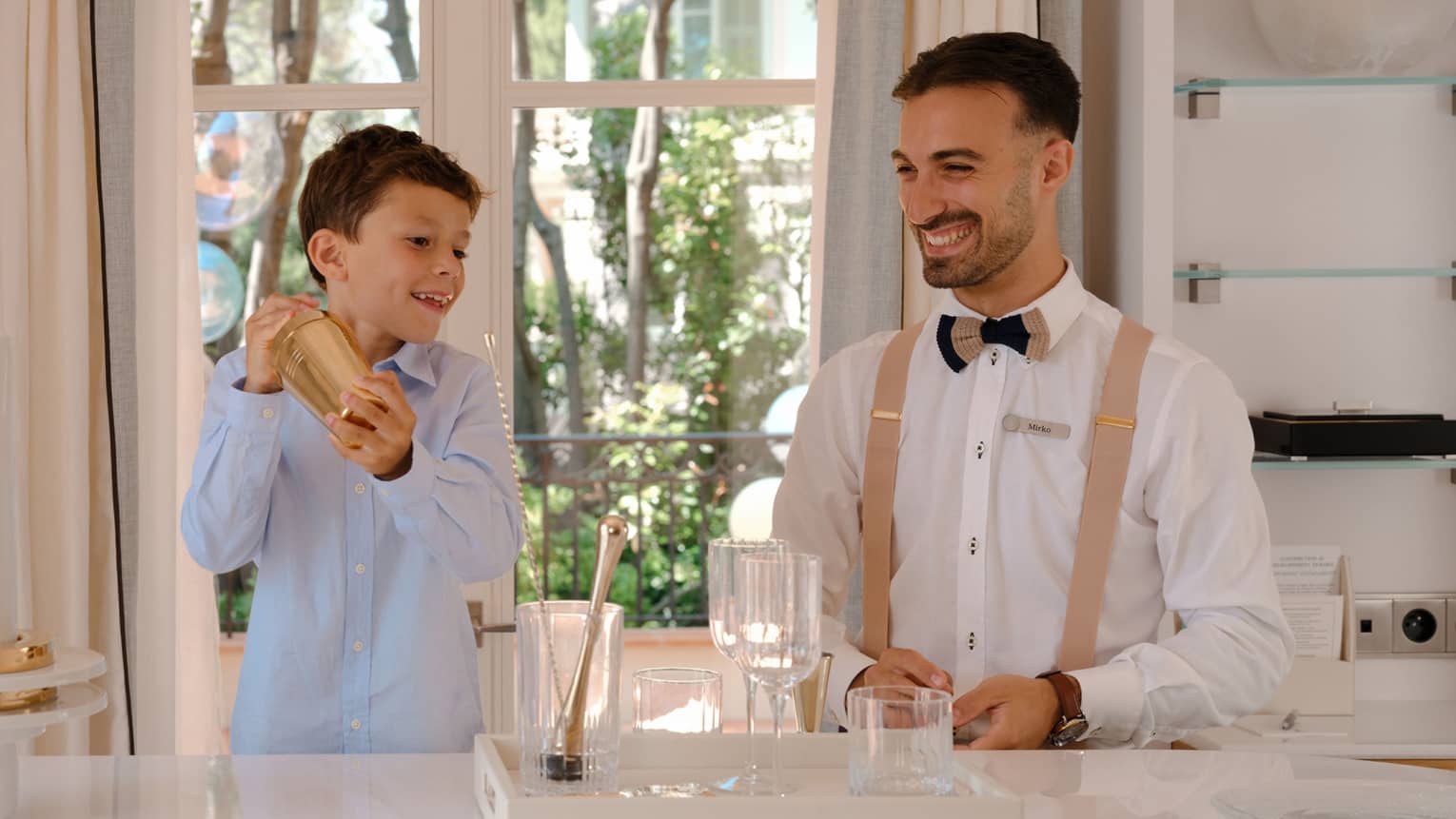 Child holds gold mocktail shaker as smiling bartender looks on